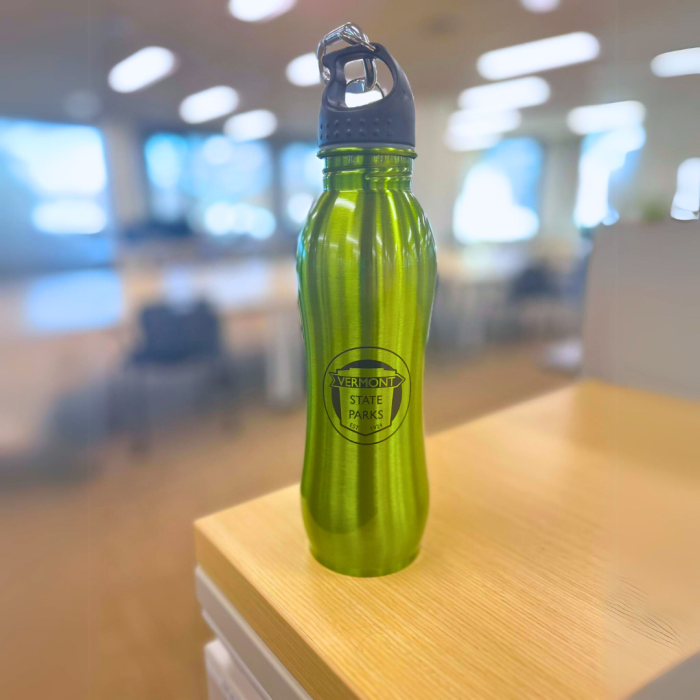 Green metal waterbottle with black shield logo sits on a desk in an office.